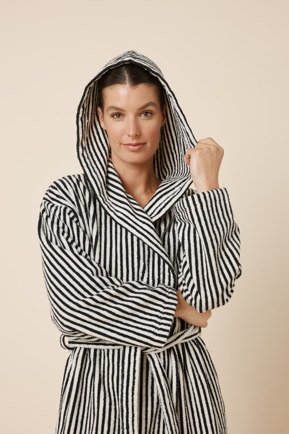 Woman wearing a striped adriana terry towel robe in black colour with the hood on holding it with her hand