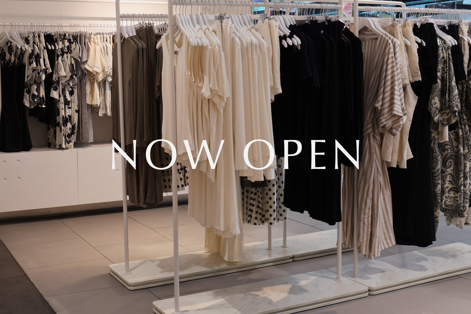 Clothing rack with various garments in a store setting, featuring 'NOW OPEN' text.