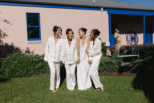 Bride in an ivory satin slip nightgown walking with three bridesmaids in matching ivory satin pyjama sets on the wedding morning