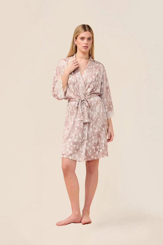 A woman modeling a light pink and white floral print silk robe with a tie waist belt, standing against a solid beige studio background.
