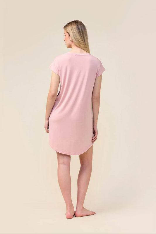 A woman standing with her back to the camera wearing a light pink short-sleeve nightshirt or tunic dress, photographed against a plain warm beige studio background.