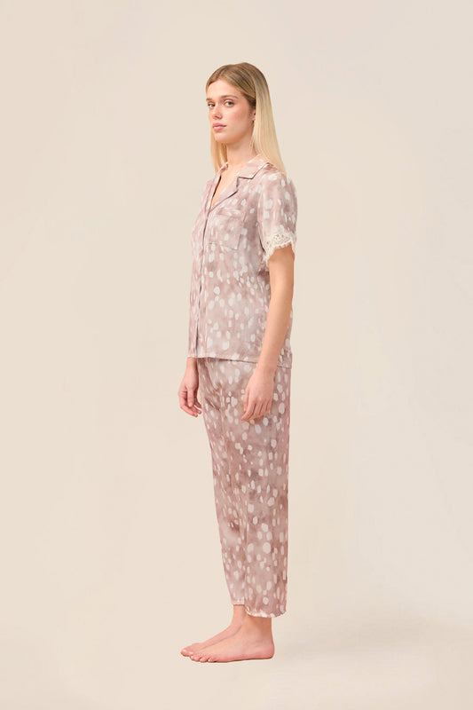 A woman stands in profile wearing a matching short-sleeve button-front pajama top and cropped pajama pants in a light blush pink fabric with a small white floral print, against a plain warm beige studio background.