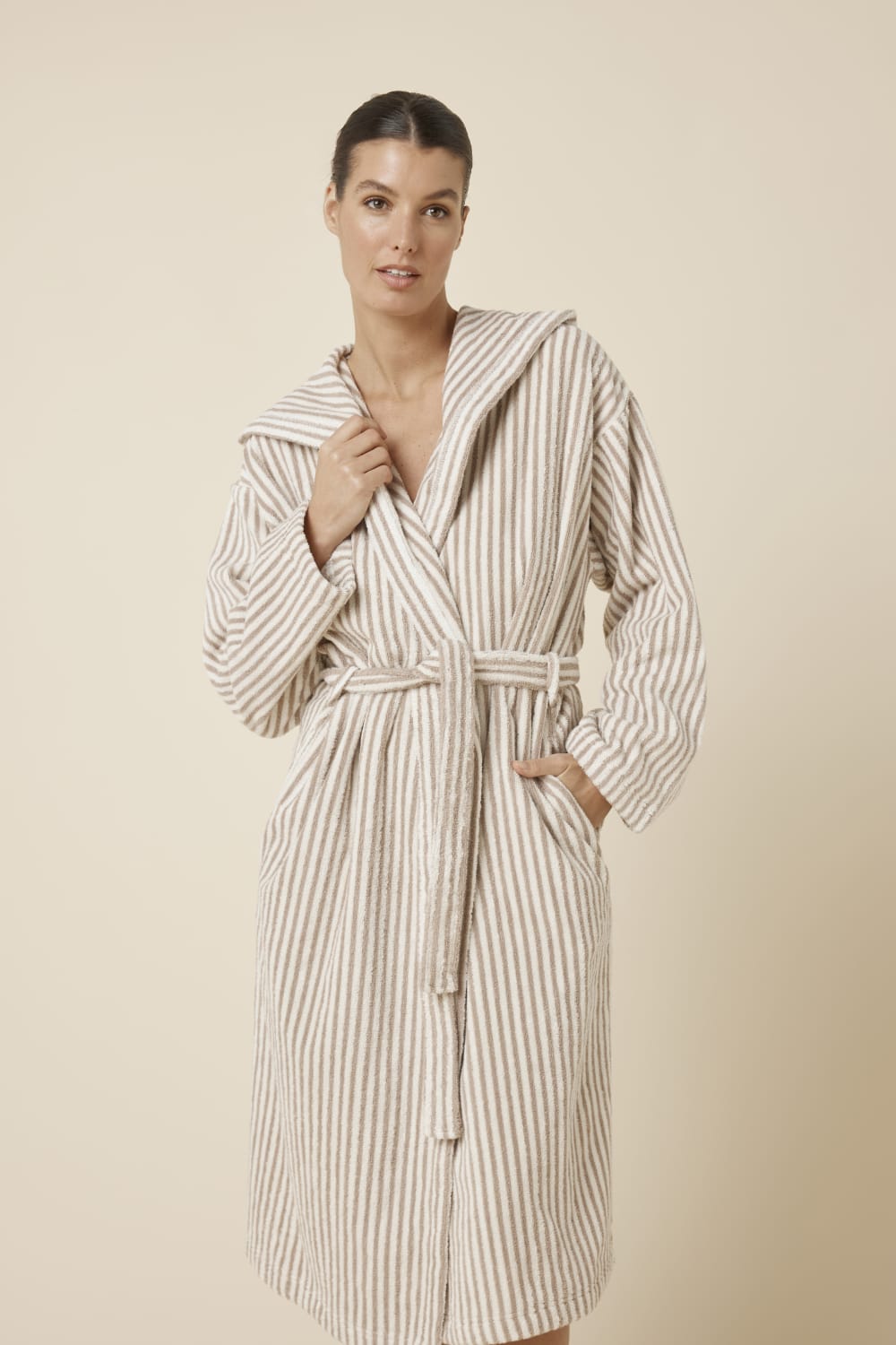 Slight close up view of woman wearing a striped adriana terry towel robe in biscuit colour standing against a plain background with hand in pocket and on upper neckline
