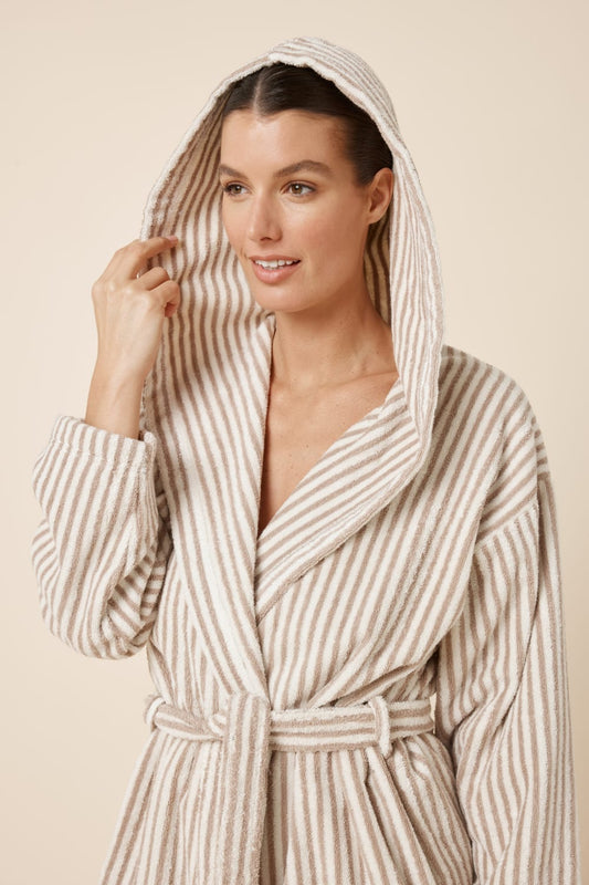 Woman wearing a striped adriana terry towel robe in biscuit colour standing against a plain background with the hood on her head