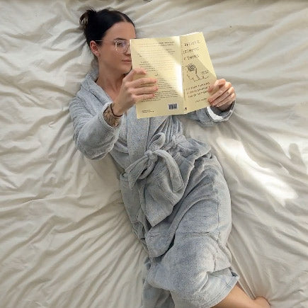 Person in a gray robe reading a book on a bed