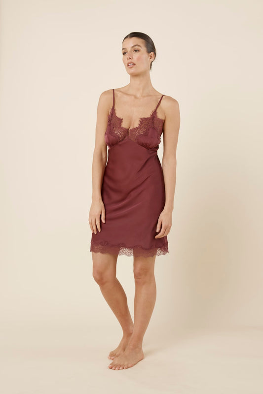 Woman wearing Azalia Silky Satin Short Chemise Burgundy standing in a relaxed pose, highlighting the lace neckline, satin slip silhouette and delicate hem trim.