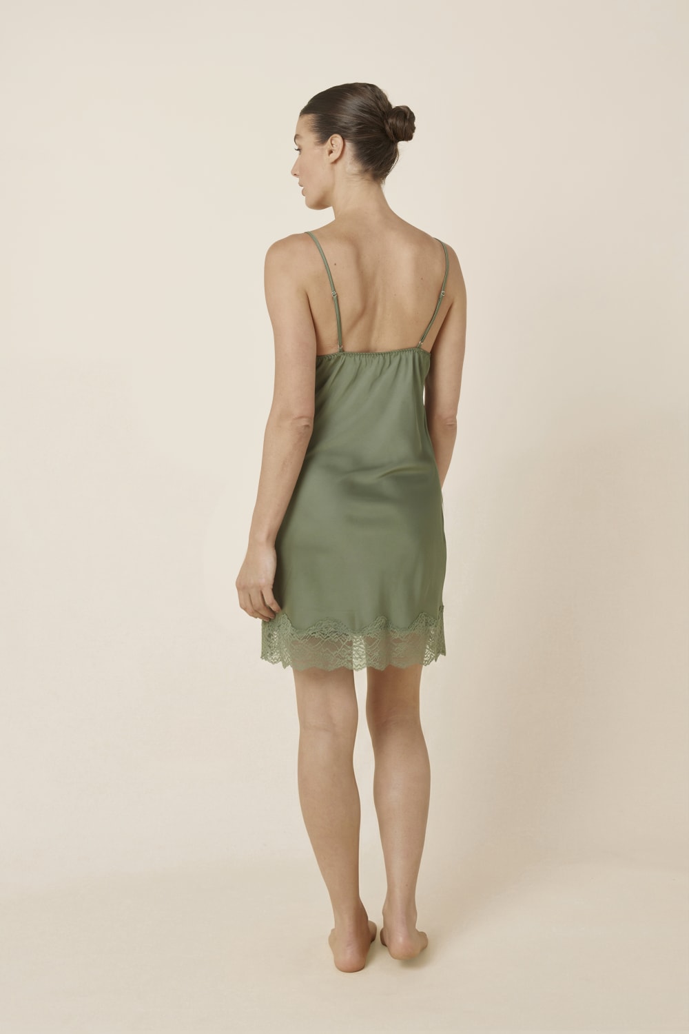 Woman wearing Azalia Silky Satin Short Chemise Olive standing turned away to show the back of the satin slip dress with slim straps and soft flowing fabric.