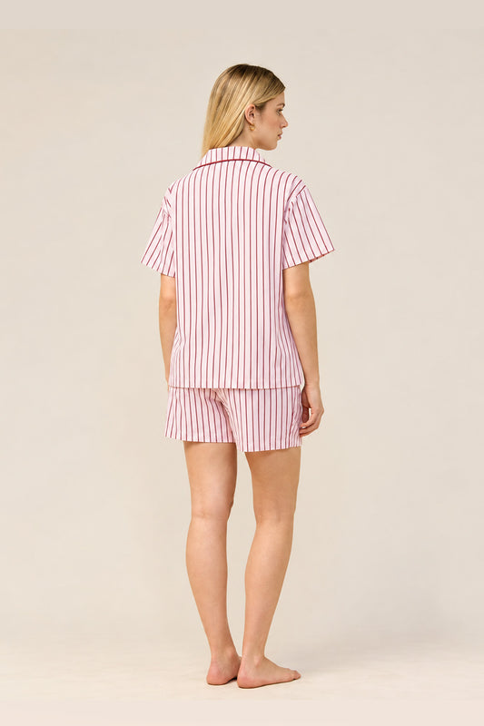 A woman stands barefoot against a plain light beige studio background, shown from the back wearing a short-sleeve collared pajama top and matching shorts with vertical pink and white stripes.