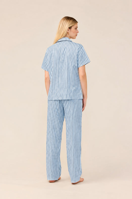 A woman stands with her back to the camera wearing a matching short-sleeve button-up pajama top and wide-leg pants set, both in a light blue and white vertical stripe pattern, against a solid beige studio background.