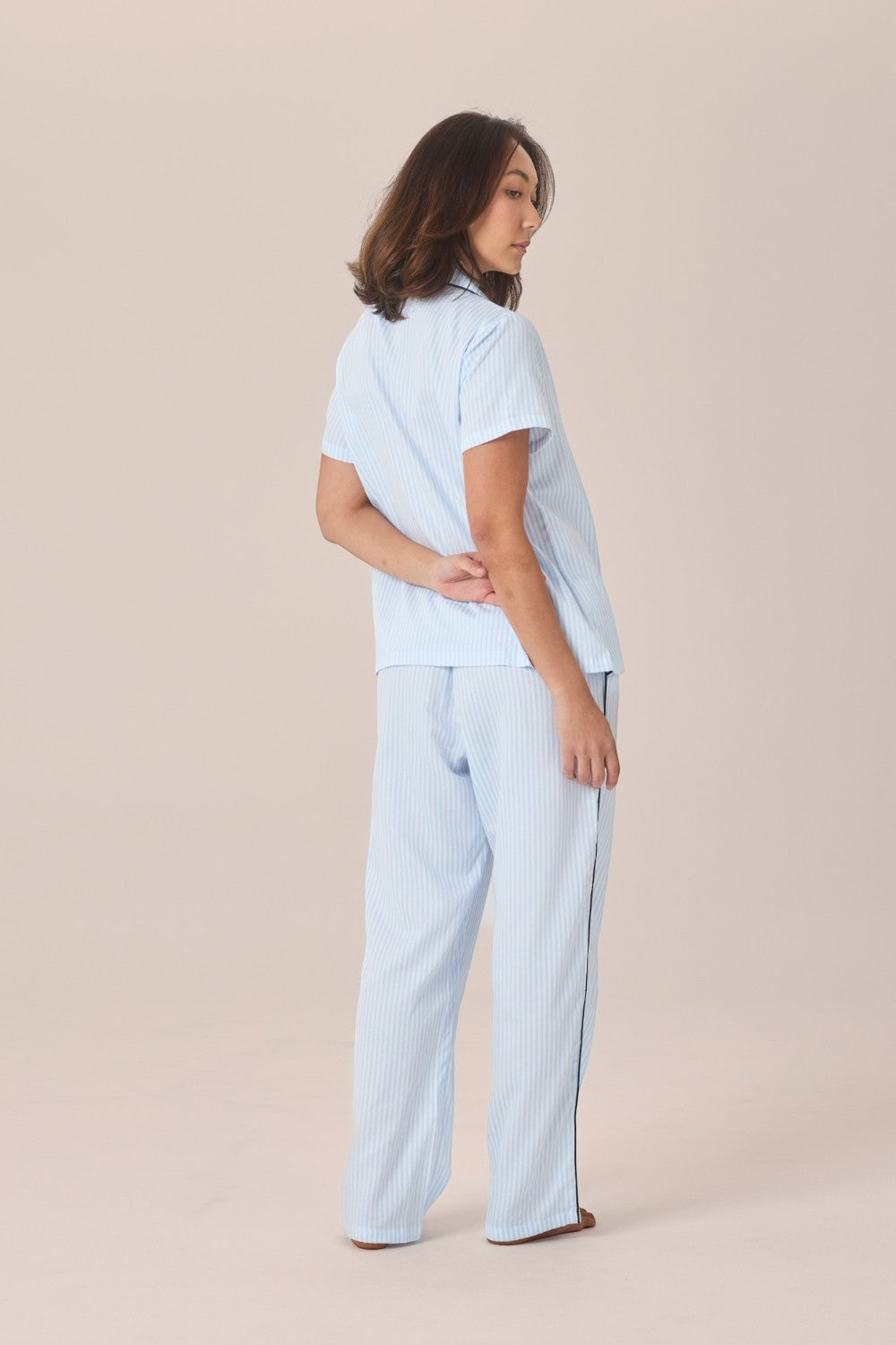 A woman viewed from behind wearing a light blue and white striped pyjama set with a short-sleeve top and long pants, standing with hands clasped behind her back.