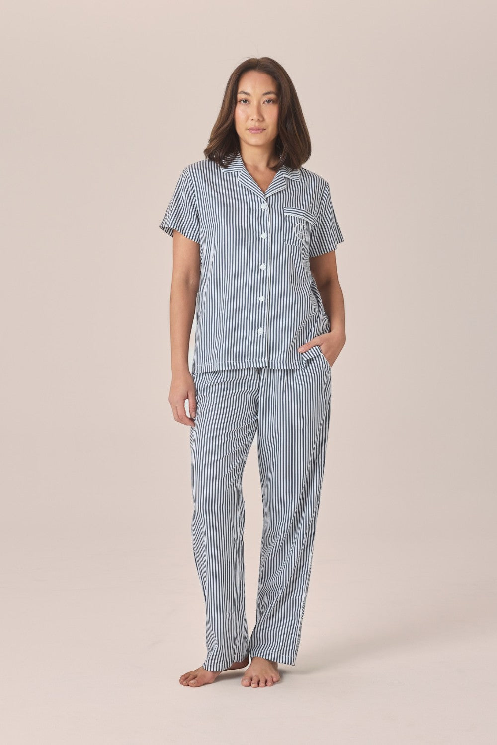 A woman modeling a navy and white striped pyjama set with a short-sleeve button-up top and long pants, standing against a solid beige studio background.