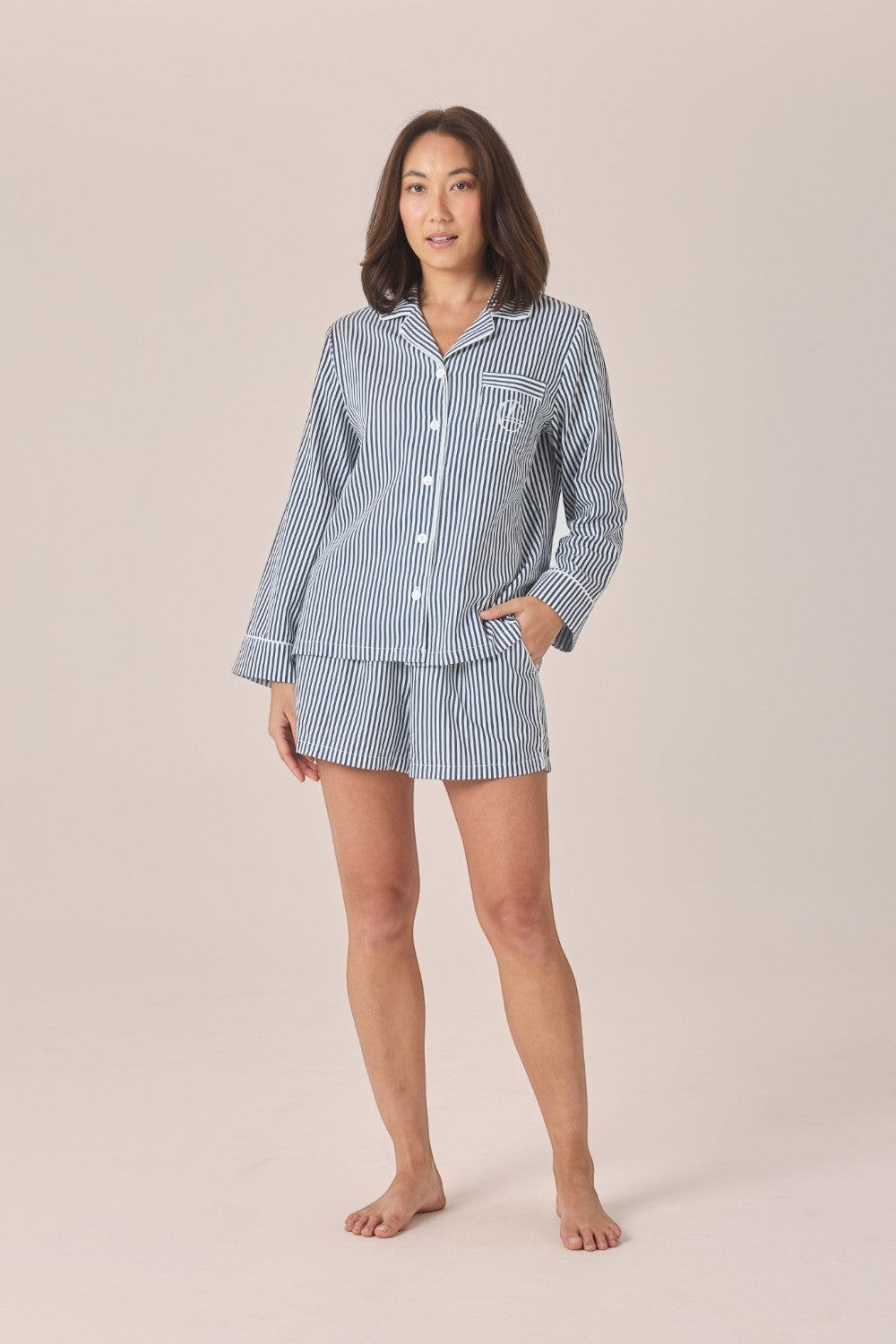 A woman modeling a matching two-piece pajama set with a long-sleeve button-front top and shorts, featuring a light blue and white vertical stripe pattern, against a solid beige studio background.