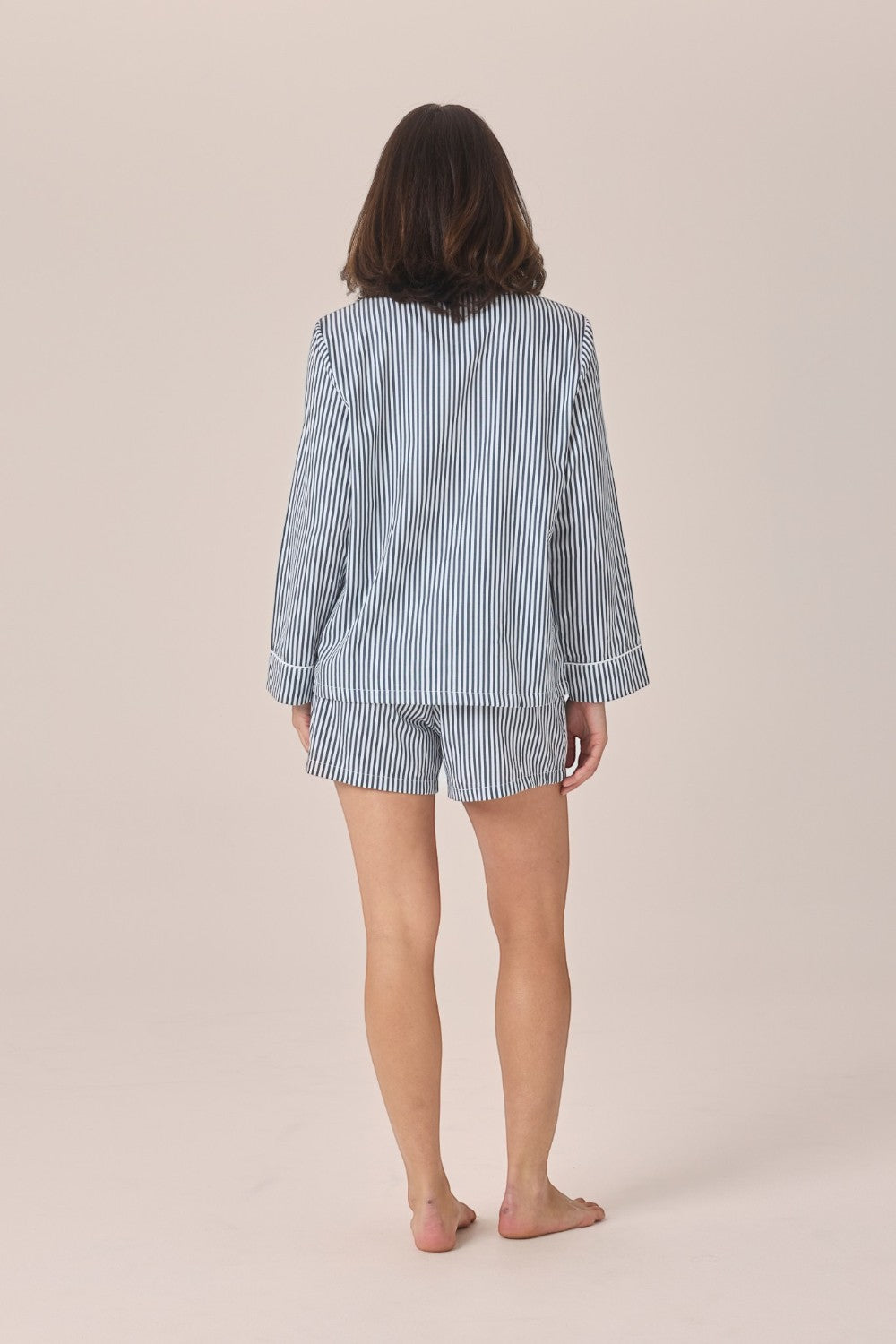 A woman viewed from behind wearing a light blue and white vertically striped pajama set with a long-sleeve top and matching shorts, standing barefoot against a plain beige studio background.