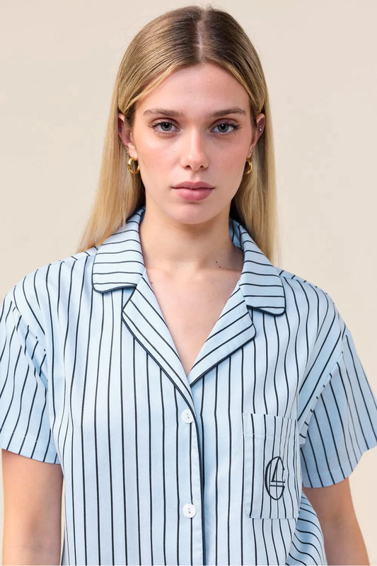 A woman wearing a light blue short-sleeve button-up shirt with thin black vertical pinstripes and a small embroidered logo on the chest pocket, set against a plain beige background.