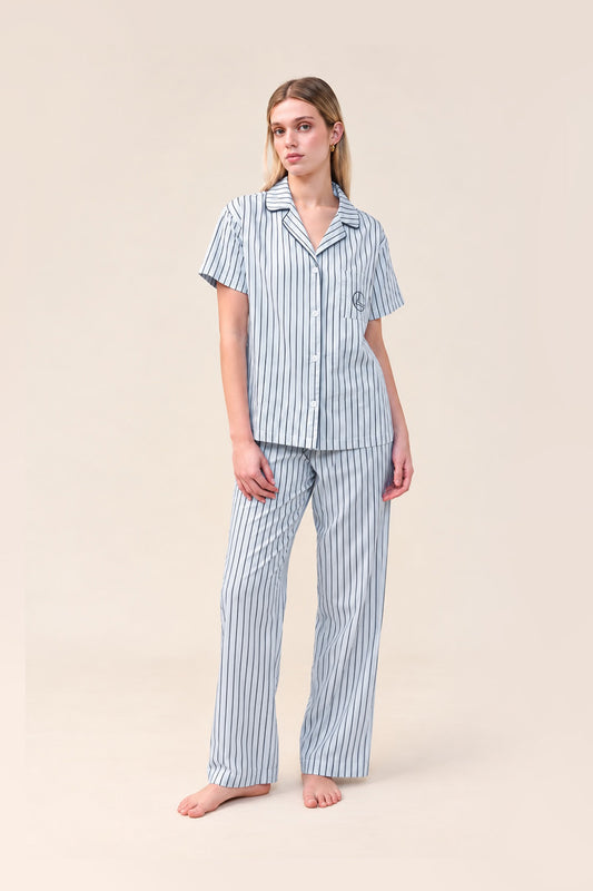 A woman modeling a matching short-sleeve button-up pajama shirt and wide-leg pajama pants set with vertical blue and white stripes, photographed against a solid light beige studio background.