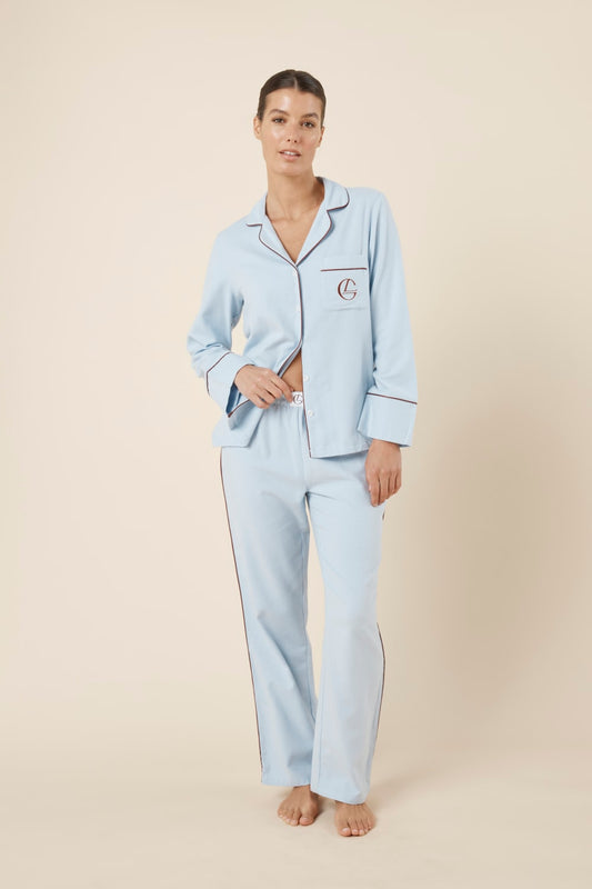 Woman wearing light blue isabella brushed cotton pyjama PJ set with a small red logo on top pocket and waistband on pant set with a beige background front on view