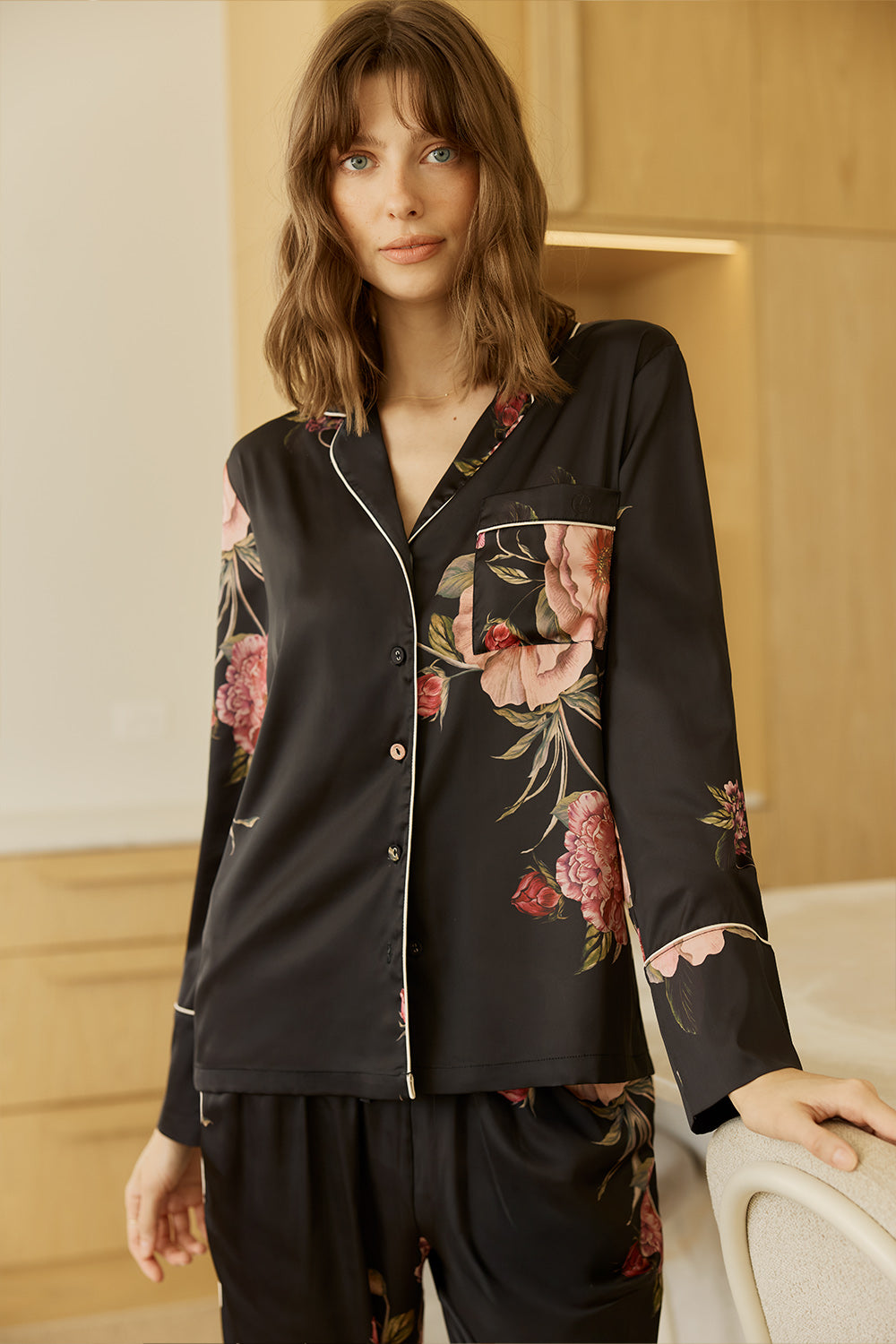 Woman wearing a Ruby Floral Satin PJ set in black with a room with wooden closet behind.