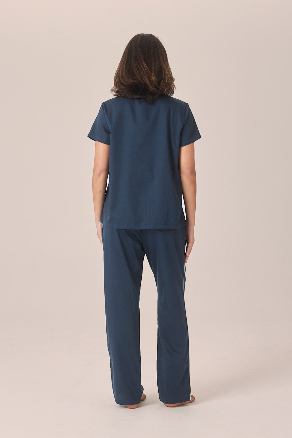 A woman viewed from behind wearing a navy pyjama set with a short-sleeve top and long pants, standing barefoot against a plain beige studio background.