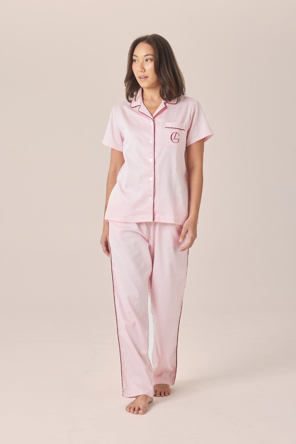 A woman modeling a matching pale pink pyjama set with a short-sleeve button-front top and long pants, featuring an embroidered logo pocket, against a solid beige studio background.
