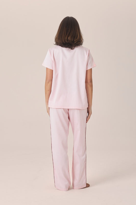 A woman viewed from behind wearing a pale pink pyjama set with a short-sleeve top and long pants, standing barefoot against a plain beige studio background.