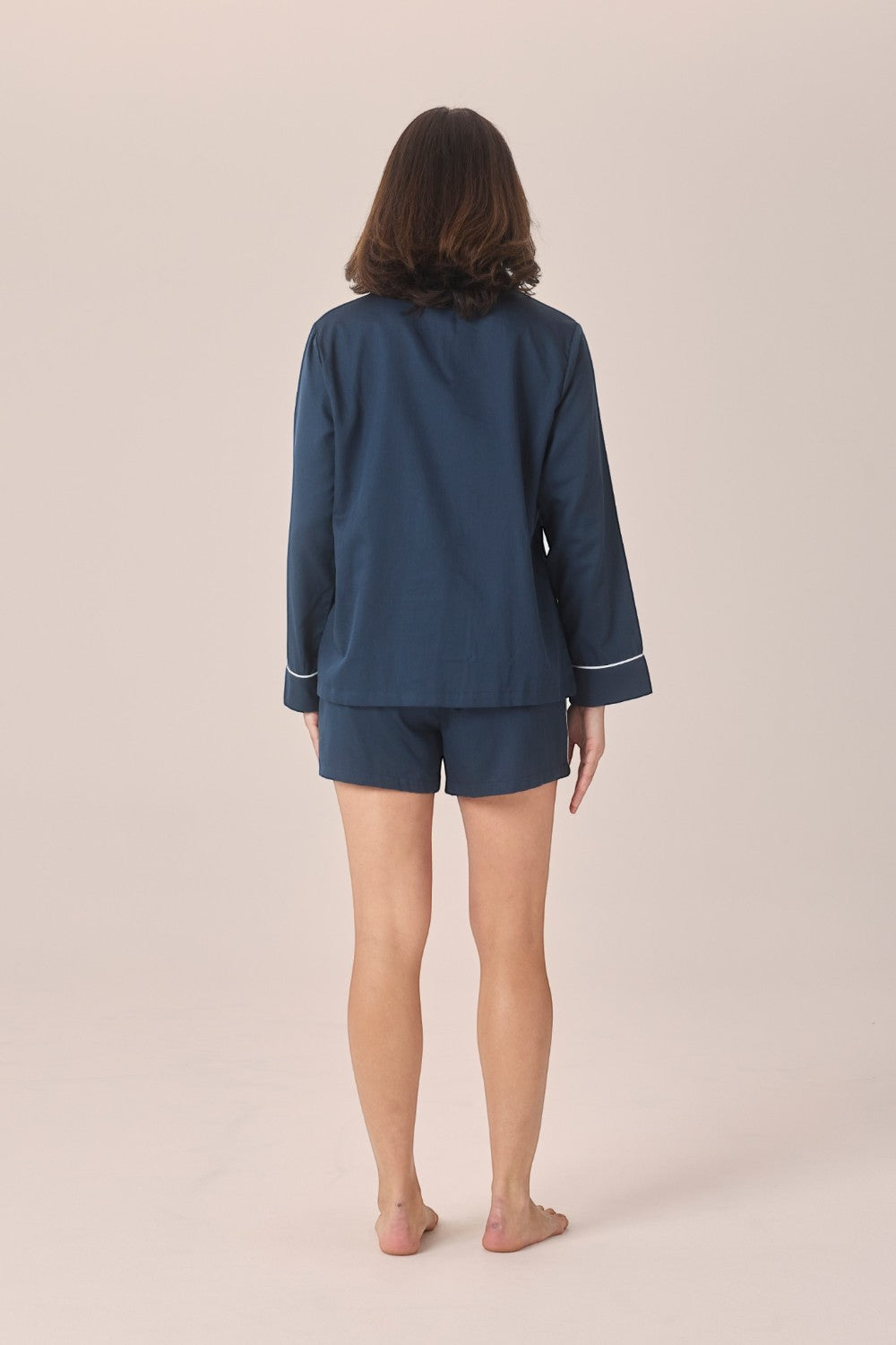 A woman stands with her back to the camera wearing a matching navy blue pajama set with white piping on the cuffs and collar, against a solid light beige background.
