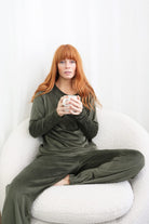 Thea Velour Long Lounge Set Olive styled on model in a relaxed seated pose wearing the long sleeve velour top and matching lounge pants holding a mug