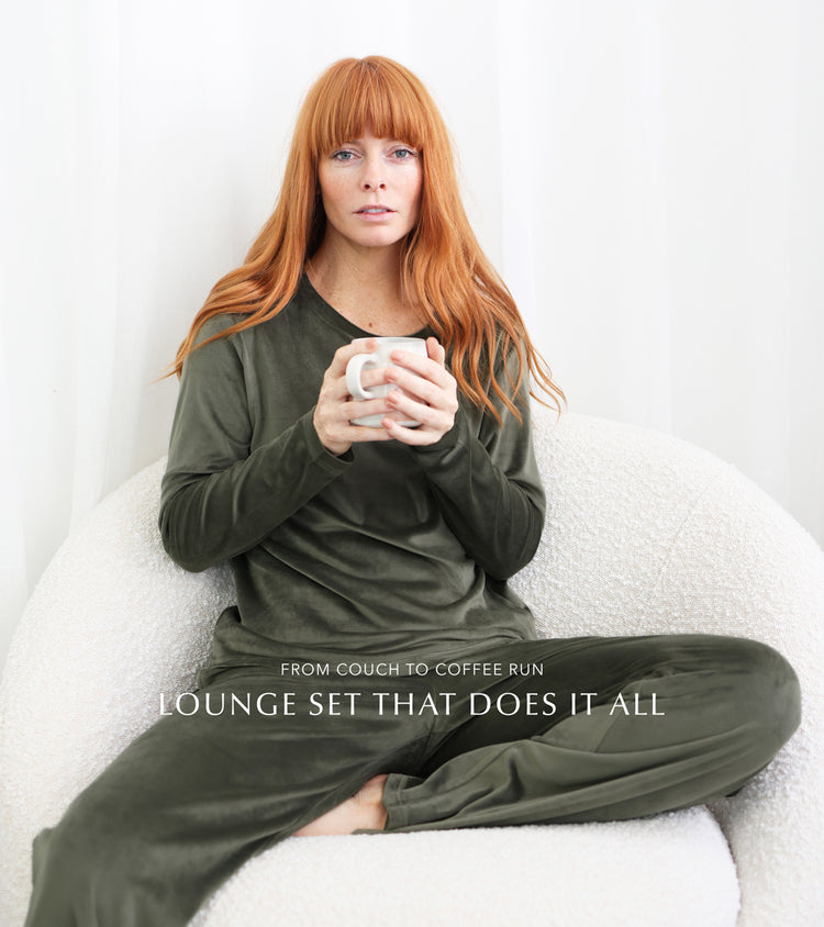 Women wearing the thea velour olive green pyjama set while holding a coffee, and lounging on a comfy sofa. 