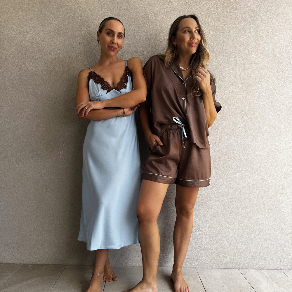Two women wearing satin pajamas standing against a neutral wall.