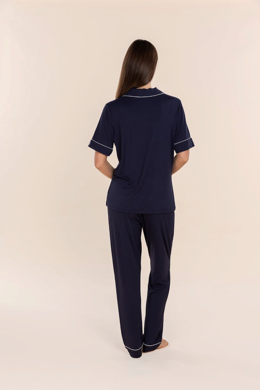 A woman viewed from the back wearing a navy blue short-sleeve pajama top and matching pants with thin white piping trim, standing against a plain beige studio background.