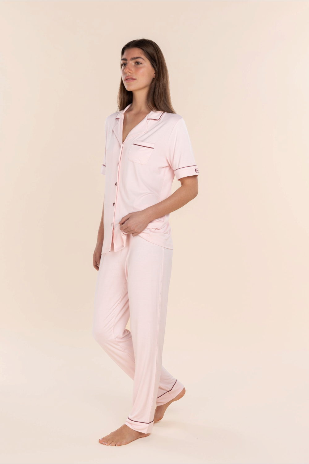 A woman modeling a light pink pajama set with thin dark pink piping details, consisting of a short-sleeve button-up shirt and matching pants, against a solid cream background.