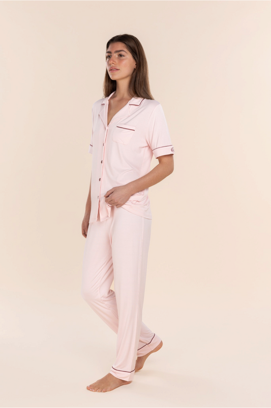 A woman modeling a light pink pajama set with thin dark pink piping details, consisting of a short-sleeve button-up shirt and matching pants, against a solid cream background.