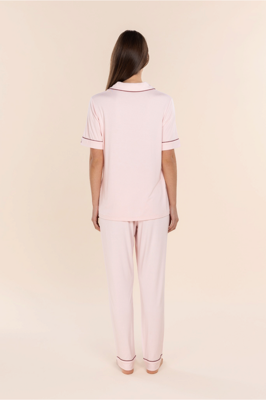 A woman viewed from the back wearing a light pink short-sleeve polo pajama set with thin dark red piping on the collar and sleeve cuffs, paired with matching light pink pajama pants, against a solid pale beige background.