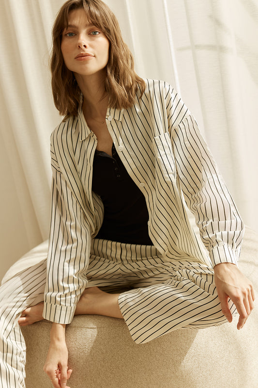 Woman wearing a black and white striped ivory willow cotton stripe pyjama PJ set in set on a curtain background sitting with black top underneath